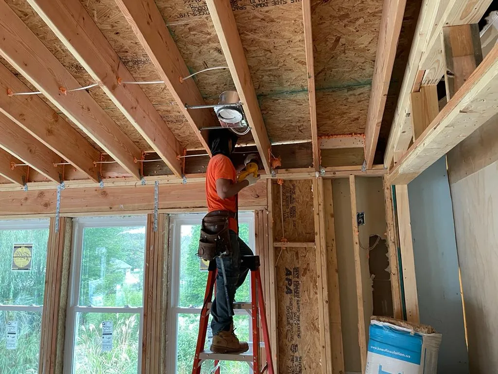 A worker on a ladder installing framing, with exposed ceiling joists and new stud walls around him.