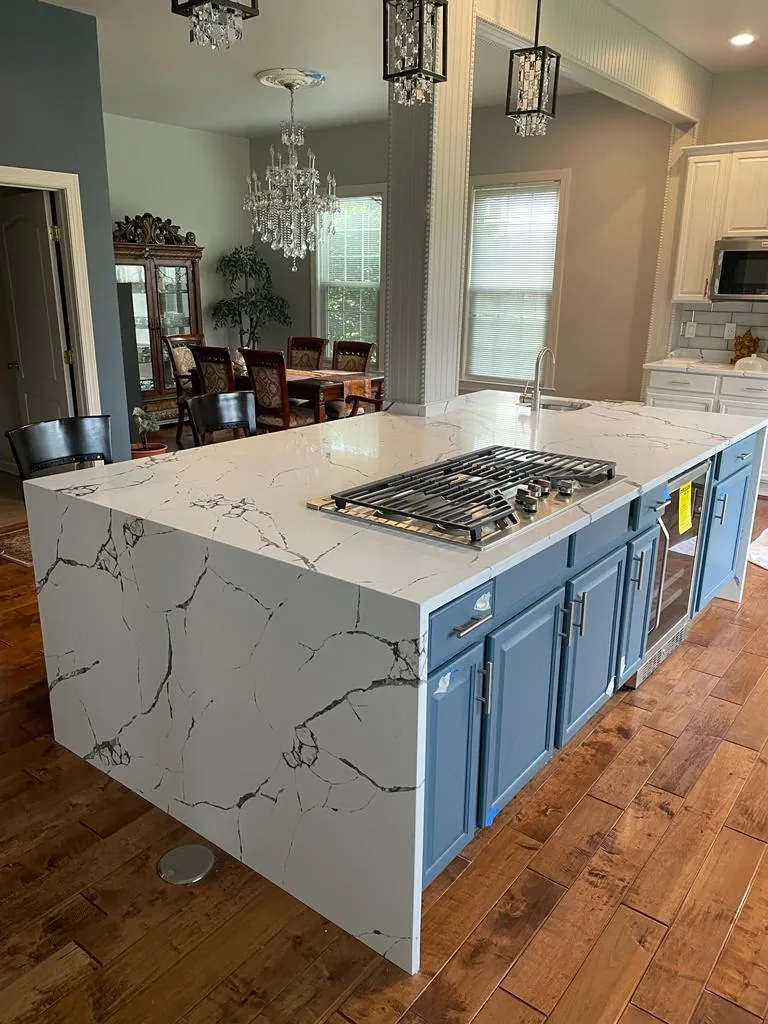 A kitchen island in blue cabinetry with a white quartz top and gas cooktop, viewed from the adjoining living room.